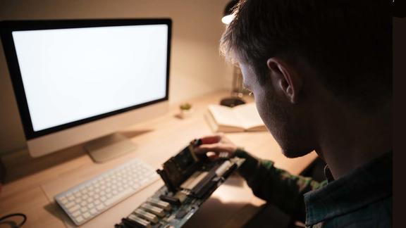 Photo: Young man repairing motherboard with blank screen computer in background.
